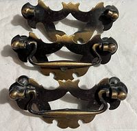 Four vintage metal drawer pulls shown stacked, displaying detailed ornate design and patina of finish.