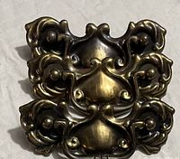 Three ornate vintage brass drawer pulls stacked on a white surface showing patina and decorative curl details.