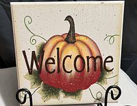 Photo shows a square plaque with a painted pumpkin and the word 'Welcome' over it, held in an easel.