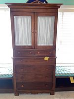 Front view of the two-piece wooden armoire showing glass paneled doors with white curtains and drawers below.