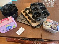Photo showing assortment of baking pans including Bundt cake pans, muffin pans, cupcake liners in a plastic container, and cookie cutters on a table.
