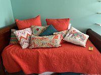 Twin sized orange quilt with detailed quilted patterns and various pillows of different colors and patterns arranged on a daybed.