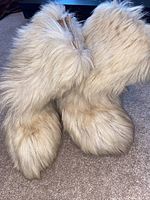 Pair of tall cream-colored genuine fur apres ski boots with thick fluffy fur and soft soles, photographed standing side by side on carpet.