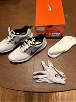 Nike Explorer SL men's golf shoes in white, black, and platinum with accompanying FootJoy golf glove and white socks laid out on surface with Nike shoebox nearby.