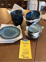 Photo showing nine stack of blue stoneware bowls, three salad-size plates, and eight small plates, all with a glossy blue finish and wrapped with packing paper.
