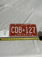 Front view of 1976 Ontario red diplomat license plate with embossed white characters CDB 127, crown symbol, and Ontario text, photographed next to a measuring tape.