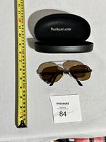 Overall view of sunglasses laid out with closed case and measuring tape showing size about 5.5 inches across.
