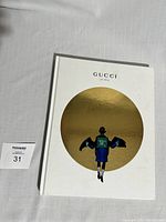 Front cover of the 2017 Gucci hardcover gift catalog book showing artwork by Ignasi Monreal—a figure with black wings against a gold circular background on a white cover.