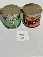 Photo showing both vintage cigarette tins side by side on a white surface with a measuring tape behind them for scale, and a lot number card in front.