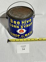 Front view of Bee Hive Golden Corn Syrup tin can showing full label, bright blue color with yellow text and beehive graphic. Tape measure indicates about 7 inches height.