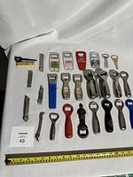 Full layout of 39 assorted beer bottle openers on white background with measuring tape for scale.