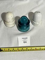 Three insulators placed on a white fabric surface with a measuring tape in the background and a labeled card (64) in front. Two are white ceramic labeled 'CPR' and one aqua blue glass insulator is in the middle.
