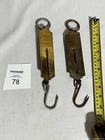 Two brass pocket balance scales with metal hooks and loop handles, placed next to a measuring tape showing about 6 inches in length.