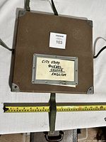 Top-down view of intact brown film reel case showing label and measuring tape at base indicating approximate size.