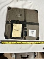 Top view of gray vintage hard carrying case with black leather straps, metal buckles, corner protection, and measuring tape placed for scale