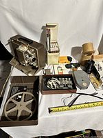 Full setup including vintage Argus Showmaster 750 projector, various film reels, accessories, cables, splicing tape, and original packaging laid out on table.