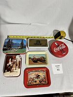 Photo showing the 6 items arranged on white textile surface with measuring tape for scale, includes 4 tins and 2 trays