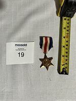 Photo showing the full medal with the attached ribbon next to a tape measure indicating about 6 cm in length.