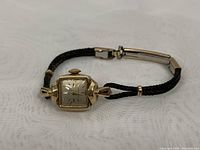 View of the 14K gold-plated ladies wristwatch with black braided leather band, square face and crystal rim, lying flat.