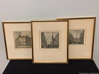 Three gold-trimmed framed etchings leaning against the wall on a black surface, showing three distinct antique city street scenes.