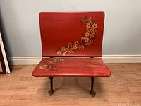 Front view of the antique wooden folding school desk converted bench showing red wooden seat and backrest with orange, white, and green floral designs. Visible wear on seat paint.