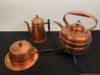 Overview of all four copper kitchenware pieces: whistling tea kettle on warmer, coffee pot, covered pot, and tray on black background