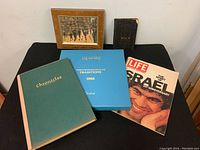 Photo of Judaica collection items on black table including framed oil painting, books, plate box, magazine, and small black book