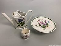 Three ceramic items including watering can with botanical illustration, pedestal cake plate with sweet pea design, and small candleholder with floral decoration