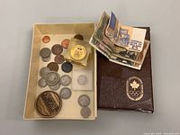 Overview photo showing a tray with various international coins including a large token, individually cased coins, and a brown leather Canadian Olympic Association case containing an Olympic medal token. Some folded banknotes are also visible.