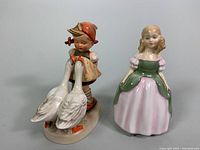 Front view of both figurines showing the Royal Doulton Penny figurine with two geese and the Goebel girl figurine standing separately.
