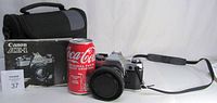 Canon AE-1 camera with lens attached, front Ricoh lens cap on, instruction booklet, camera bag, and Coca-Cola can for scale.