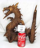 Angled front view of the wooden dragon sculpture with a soda can for size comparison. Shows detailed carving of scales, face, and claws.