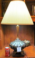 Front angle view of the full lamp showing the ceramic base, wooden accent, pleated fabric lampshade, and metal feet. A soda can is present for size reference.