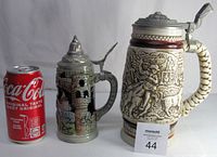 Two stoneware beer steins, one Avon cowboy-themed by Ceramarte measuring 9.25" and one Western Germany stein with castle decoration measuring 7.25". Both have lids, one a white metal lid, the other pewter.