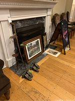 Three metal candle holders of different heights, framed prints and music sheet leaning on the fireplace, black sandwich board easel nearby, woody floor.
