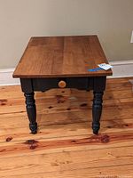 Front view of wood end table showing natural wood top, black turned legs, single drawer with knob, and wood floor background.