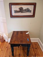 End table with stained wood top and black legs with a single drawer, placed on wooden floor against wall