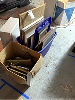 Two large boxes filled with assorted picture frames and some pictures, frames predominantly wooden in various sizes and colors, with some metal frames.