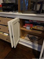 White wooden cabinet with open door showing board games inside including Monopoly, Ouija, Blokus, CSI, Imaginiff, FACT or TRAP, and a white dice shaker.