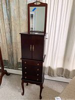 Front and side views of tall dark wood jewelry cabinet with mirror against curtain background