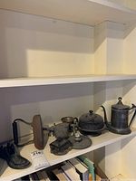 Shelf display of multiple metal items including the boot door stop, candlesticks, trivet, baby cup, and tankard