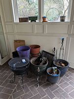 Wide shot showing grouped planters, plant stands, garden trellis, framed panels, and gardening items clustered on a porch floor near a window sill with additional small pots.