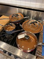 Six frying pans and saucepans with copper-colored interior and stainless steel handles, each paired with a glass lid with black knob, arranged on stove top and counter.