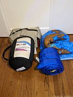 Eddie Bauer sleeping bag tote bag and two additional sleeping bags including Scooby-Doo theme, and a rolled blue memory foam topper on wooden floor near the wall.