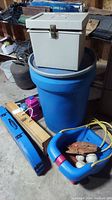 General overview photo showing blue plastic barrel with gray metal file box on top, wooden horseshoe set, blue badminton case, and blue plastic child swing holding baseball glove and balls.
