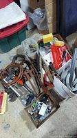 Wide angle photo of assorted clamps, copper pipes, hand tools, brush, and boxes with small hardware parts in a basement or workshop setting.