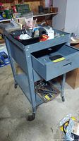 Overall view of gray metal cabinet/bench on wheels with drawer open showing tools inside. Top tray holds miscellaneous tools and parts.