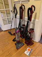 Three Shark upright vacuums standing on floor near door, showing red and gray colors and multiple attachments.