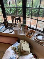 Photo shows assortment of musical note wall decor on window ledge, white ceramic plant pots, small animal figurines in wicker basket, holiday themed plates stacked, folded green cloth napkins, decorative ceramic bottle, and garden fertilizer spikes.