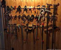 Full view of assorted clamps hanging on wooden wall including bar clamps, corner clamps, and C clamps in varied sizes and colors